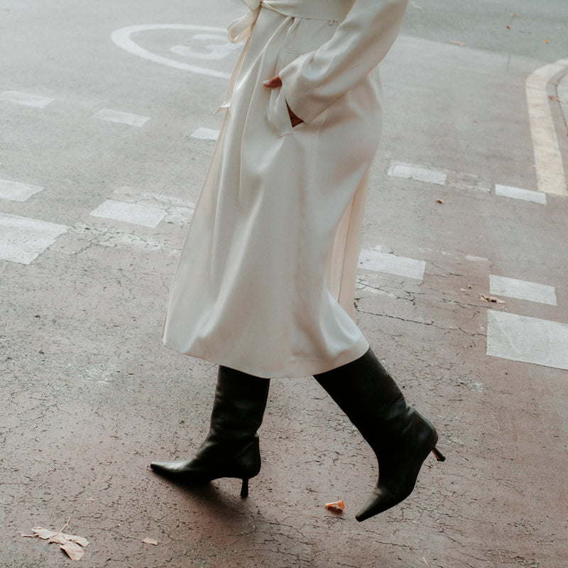 Chica caminando con unas botas de mujer en piel negra y un abrigo blanco
