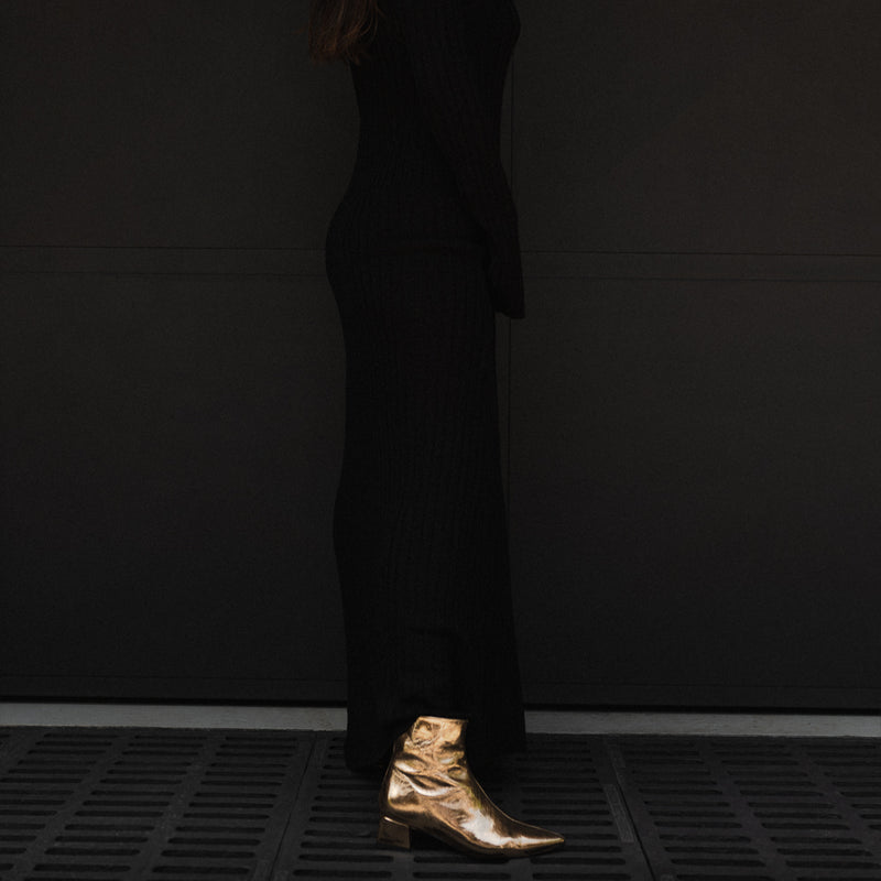 Mujer con vestido largo negro de perfil con unos botines dorados a juego